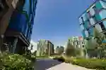 Four tall modern buildings set back against a light brick path with trees and hedges either side with a clear blue sky.