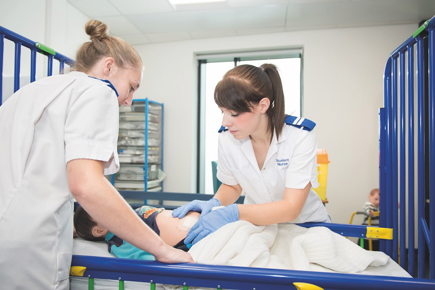 Two student nurses in white uniforms care for a young patient