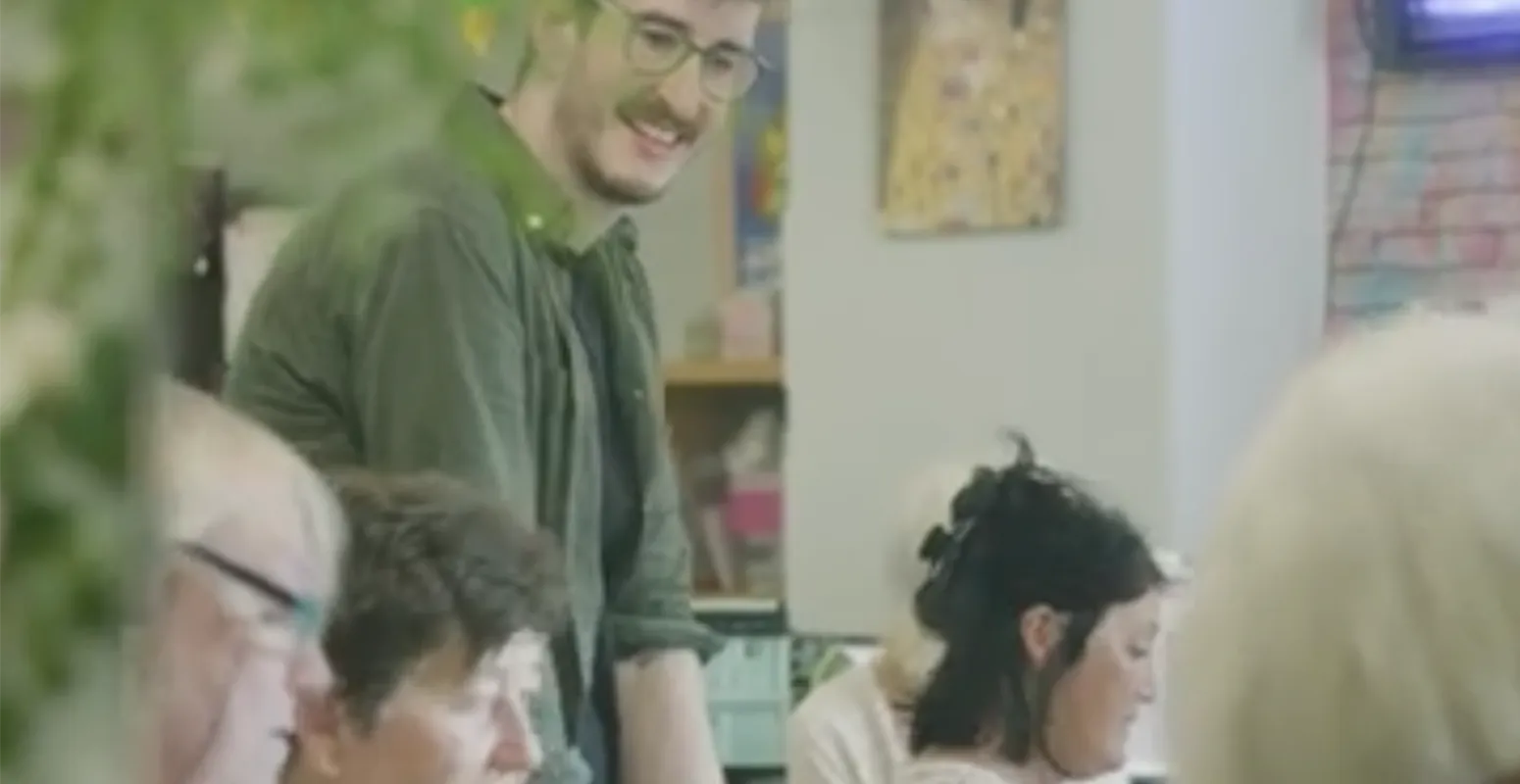 Young man with glasses stands and chats to a group of older ladies in a classroom setting