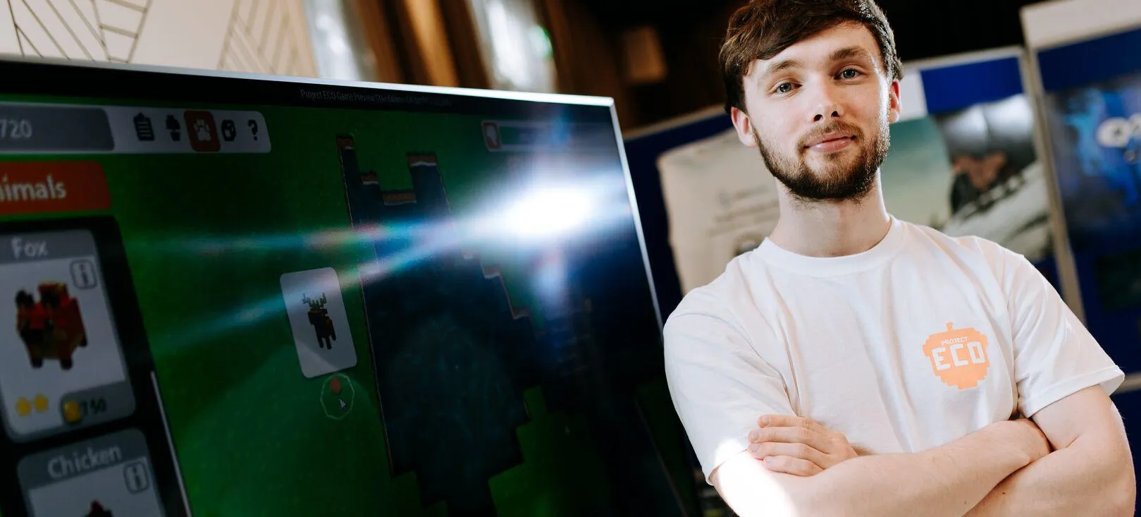A male Game Design student in a white t-shirt standing in front of a large computer screen