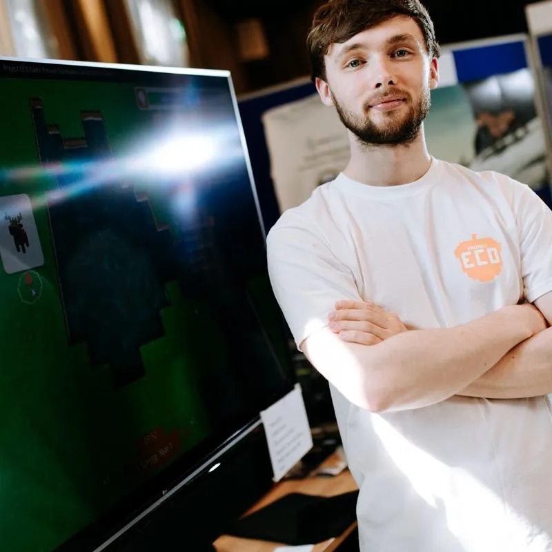 A male Game Design student in a white t-shirt standing in front of a large computer screen