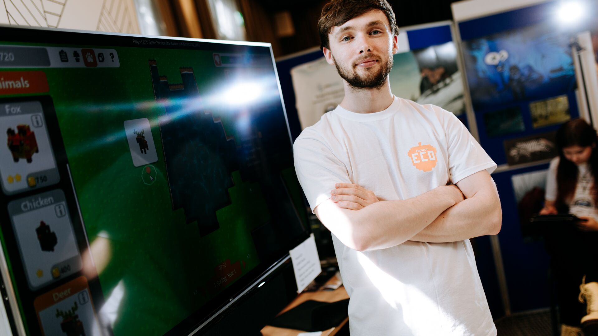 A male Game Design student in a white t-shirt standing in front of a large computer screen