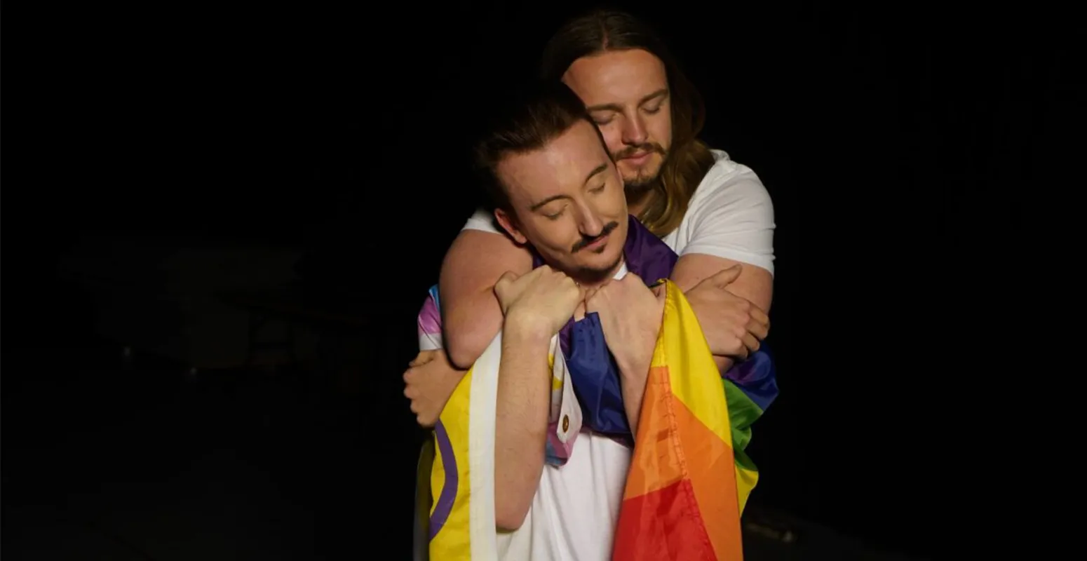 Two members of the Safer cast, embracing and holding a pride flag