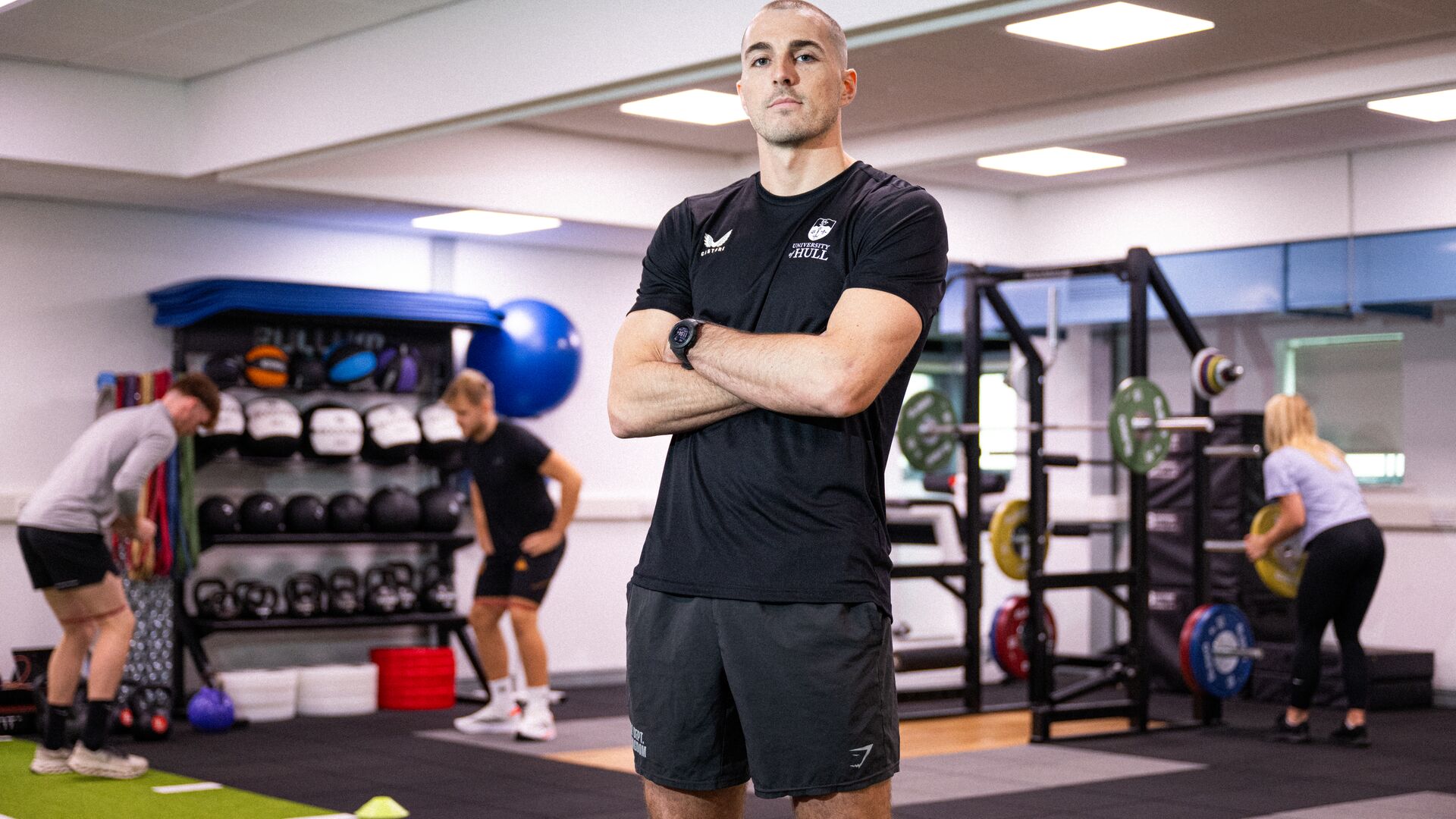 Sports student Danny Uscroft-Mould stands, arms crossed, in a gym while other users workout on equipment behind him