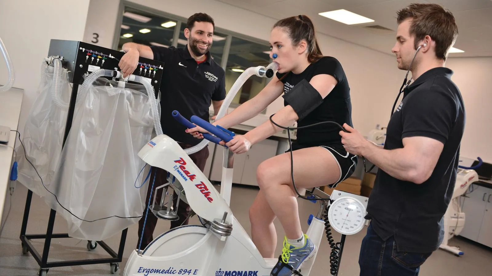 Three students in a gym, one on an exercise bike with a breathing tube on, the other two are monitoring her progress