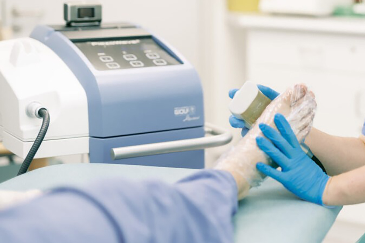 A clinician in blue gloves holds a shockwave device against a patient's foot