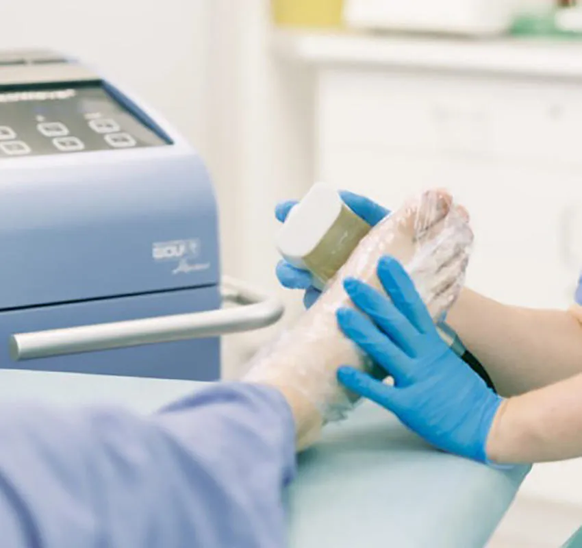 A clinician in blue gloves holds a shockwave device against a patient's foot