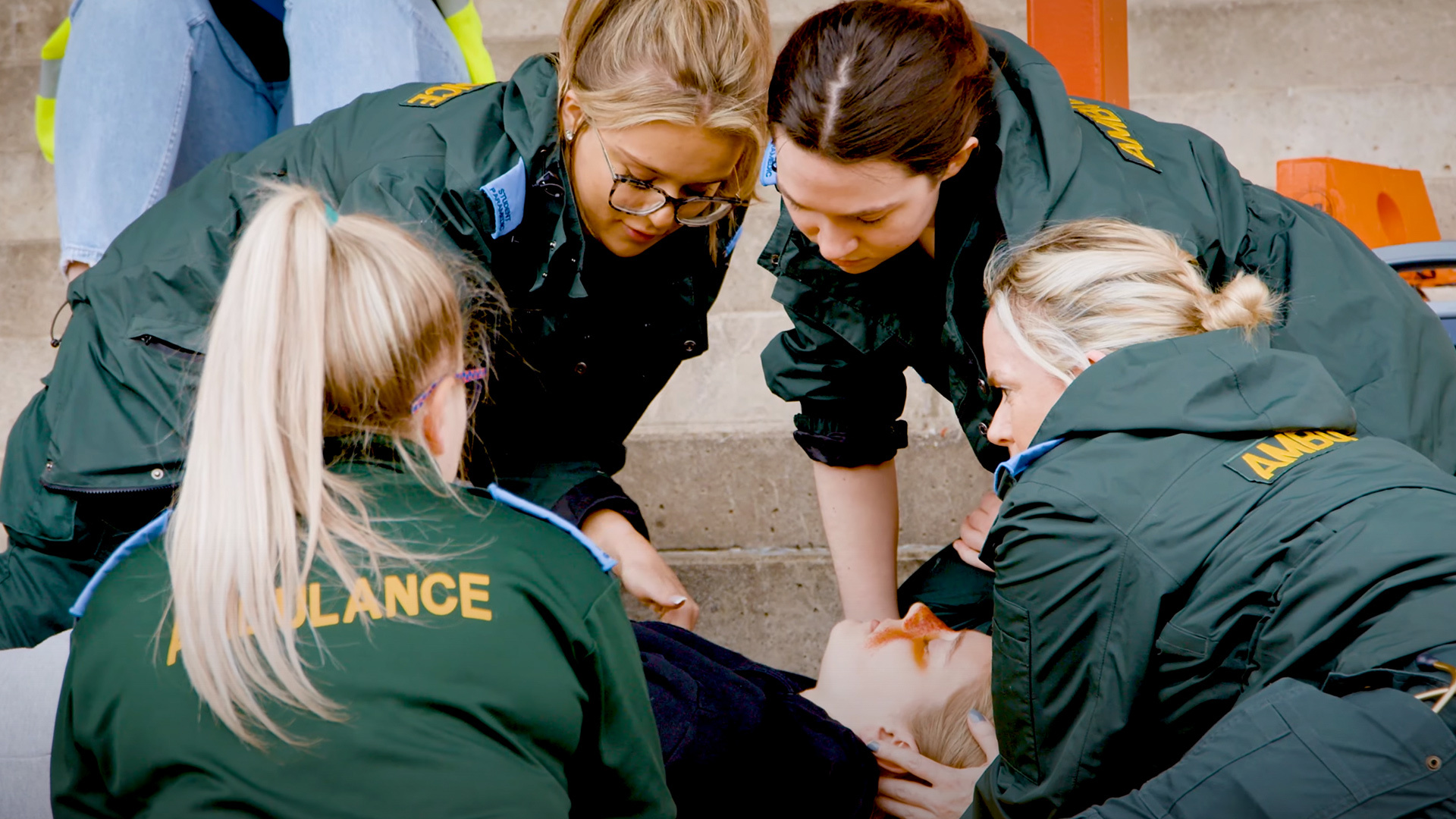 Four student paramedics in green uniforms surround a patient as part of a training exercise