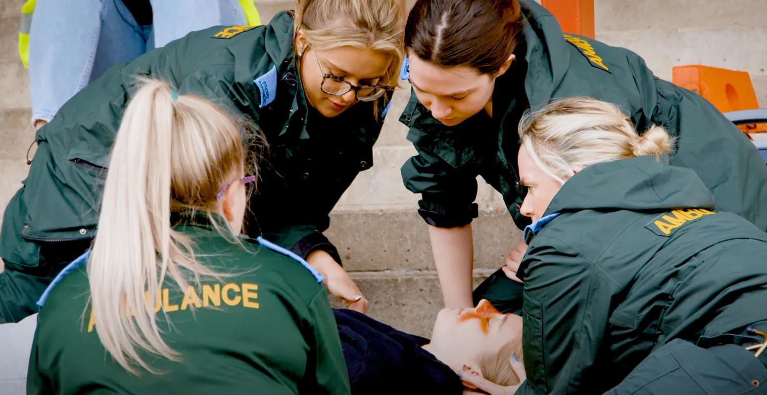 Four student paramedics in green uniforms surround a patient as part of a training exercise