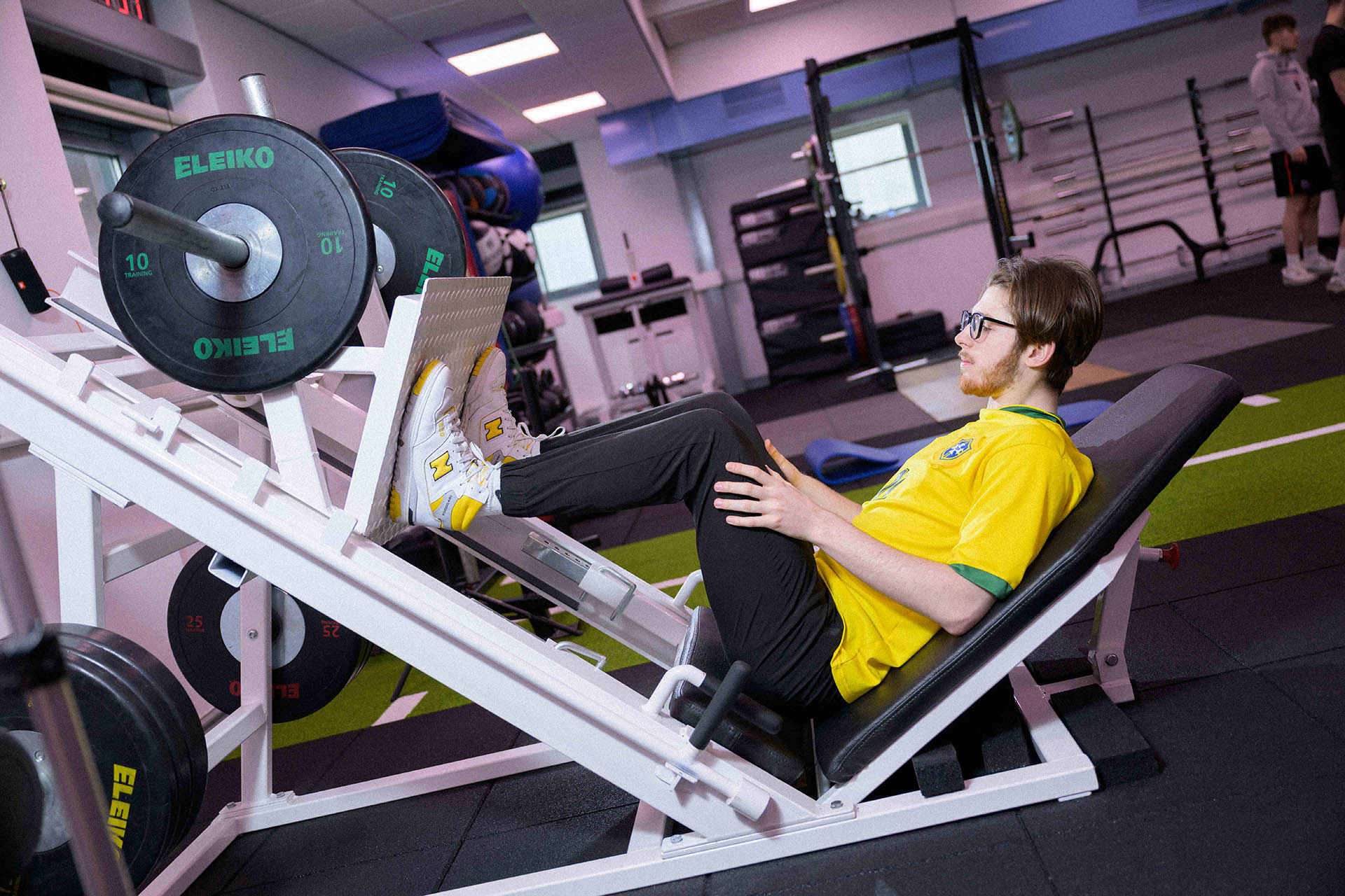 A man wearing a yellow shirt sits on a bench inside a gym surrounded by gym equipment