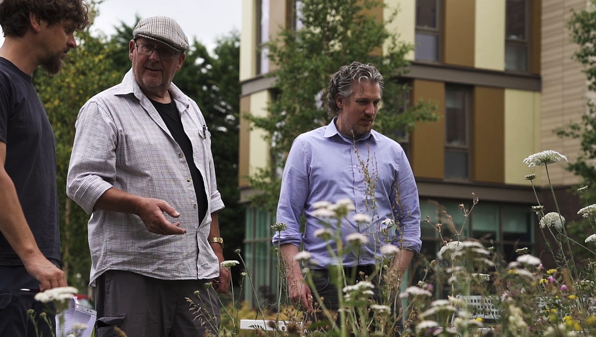 Three men stand in front of a building adorned with colorful flowers, smiling and engaged in conversation.