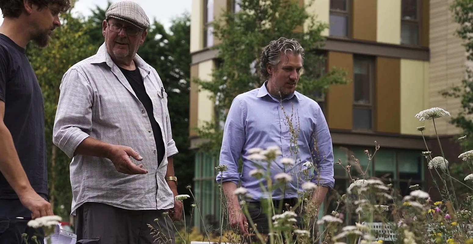 Three men stand in front of a building adorned with colorful flowers, smiling and engaged in conversation.