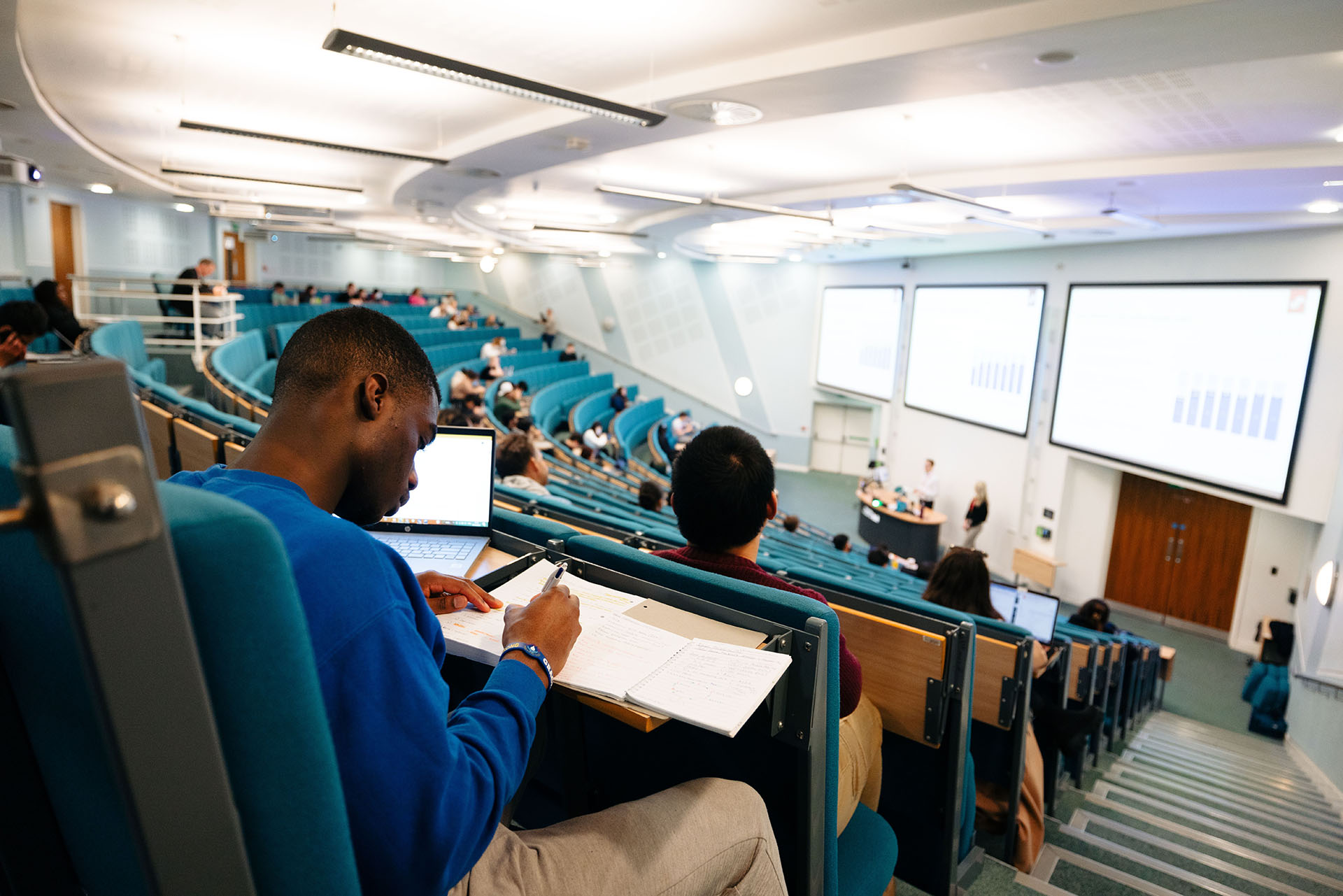 A young male student sat at the back of a lecture theatre writing with other students around him.