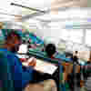 A young male student sat at the back of a lecture theatre writing with other students around him.