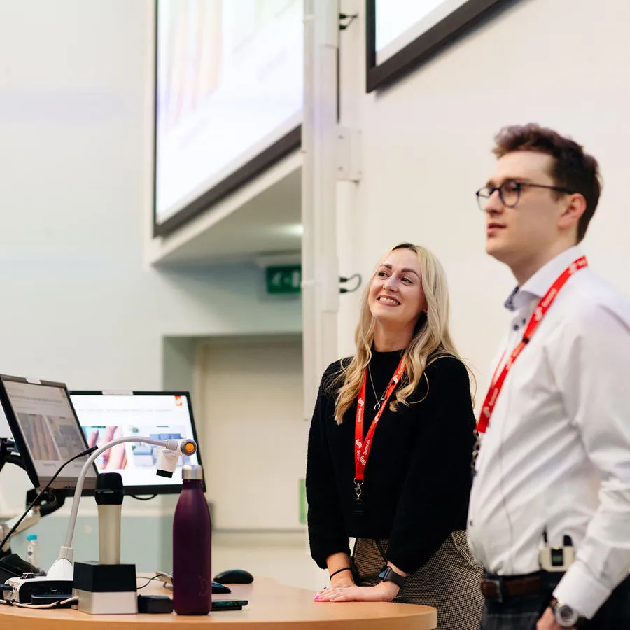 Woman and a man stood at the front of a lecture theatre with screens behind