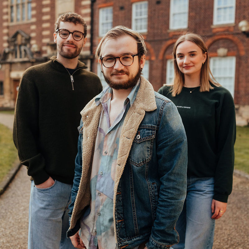 Two males and a female, all in casual clothing stand on a gravel path in front of a red-brick historic building.