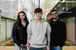 Three students stand and smile confidently in front of a window inside the Wilberforce building