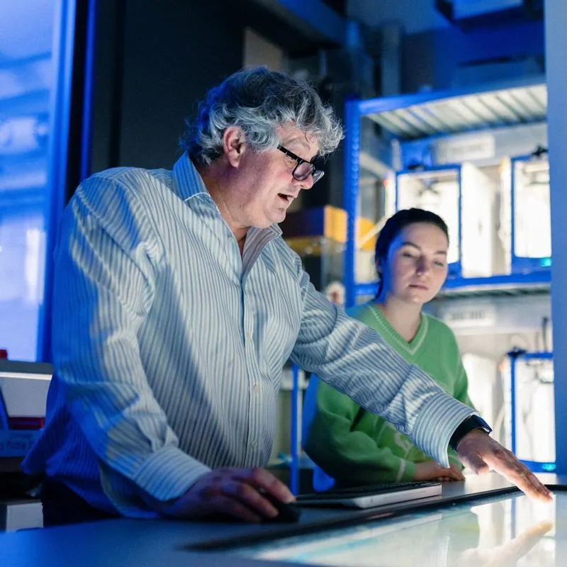 A lecturer gestures towards an item on a screen embedded in a table, surrounded by students in an engineering lab
