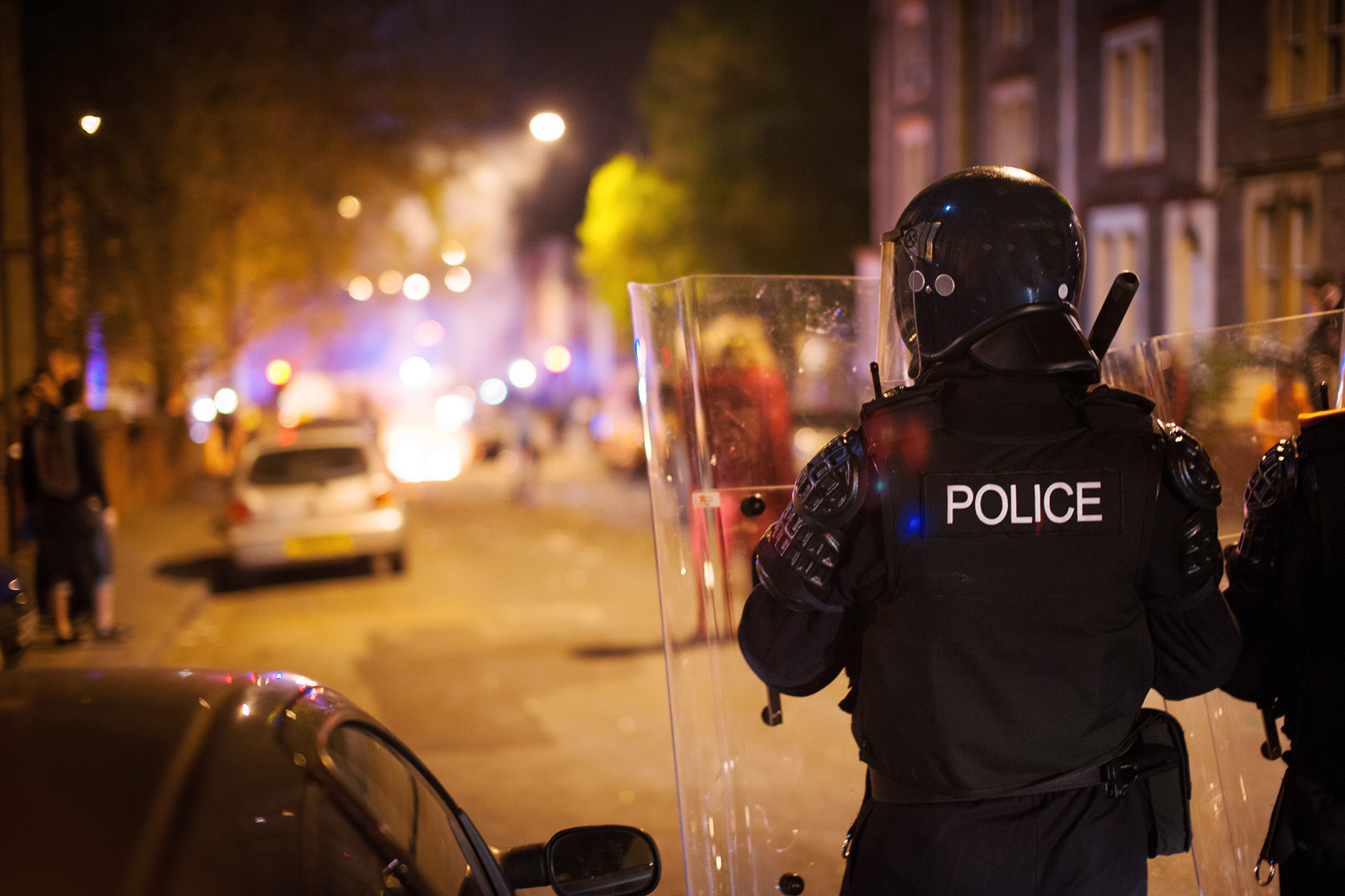 Riot police officers guarding a street at night while a fire burns ahead. 