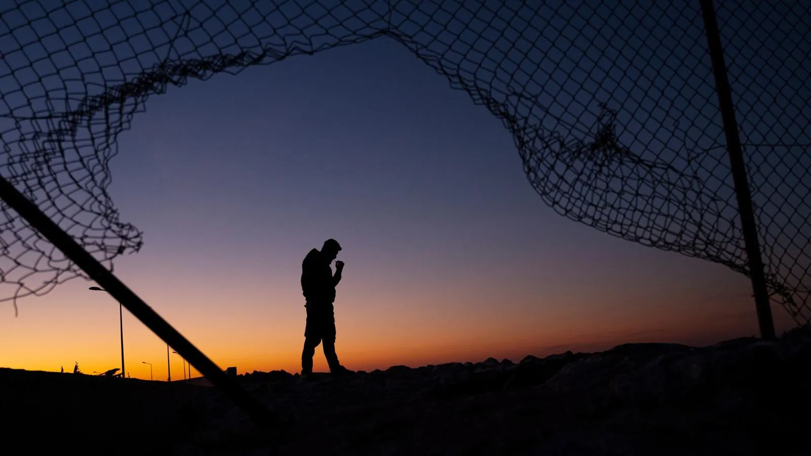 Refugee Man stood behind Broken Fence at Sunrise