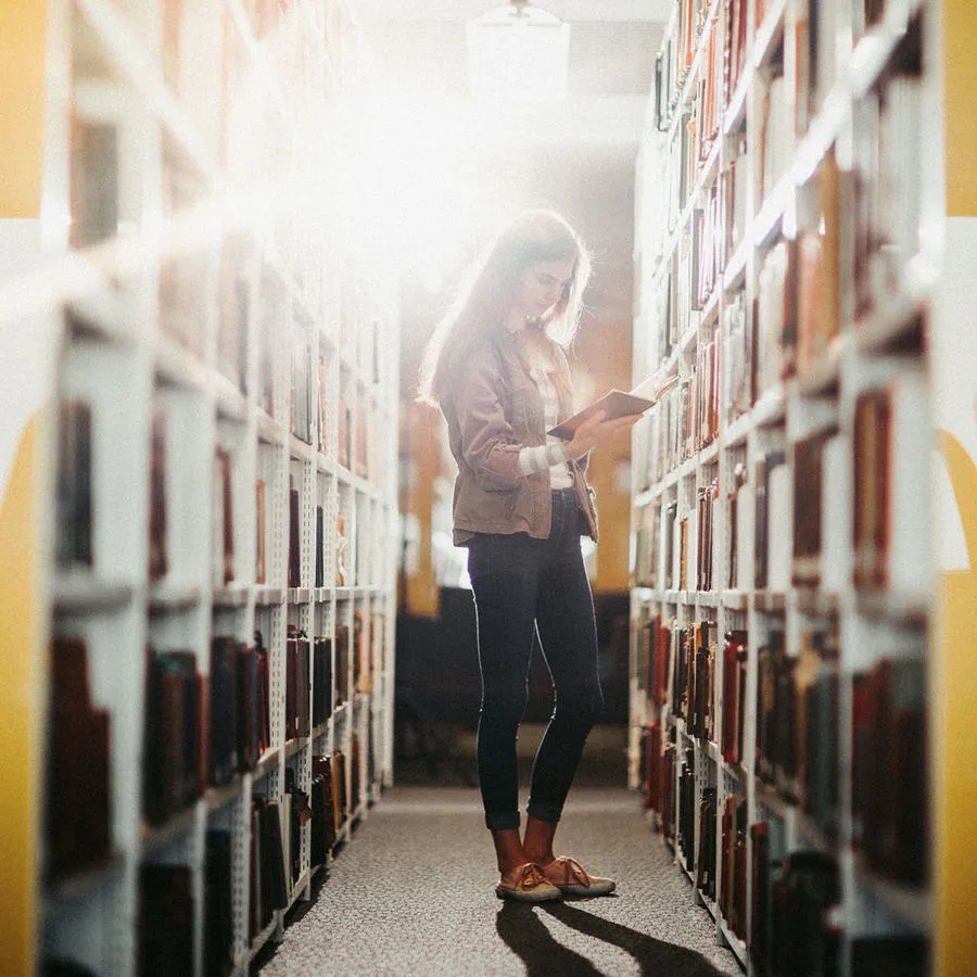 A young woman reads a book with tall shelves of books on both sides in a brightly lit room