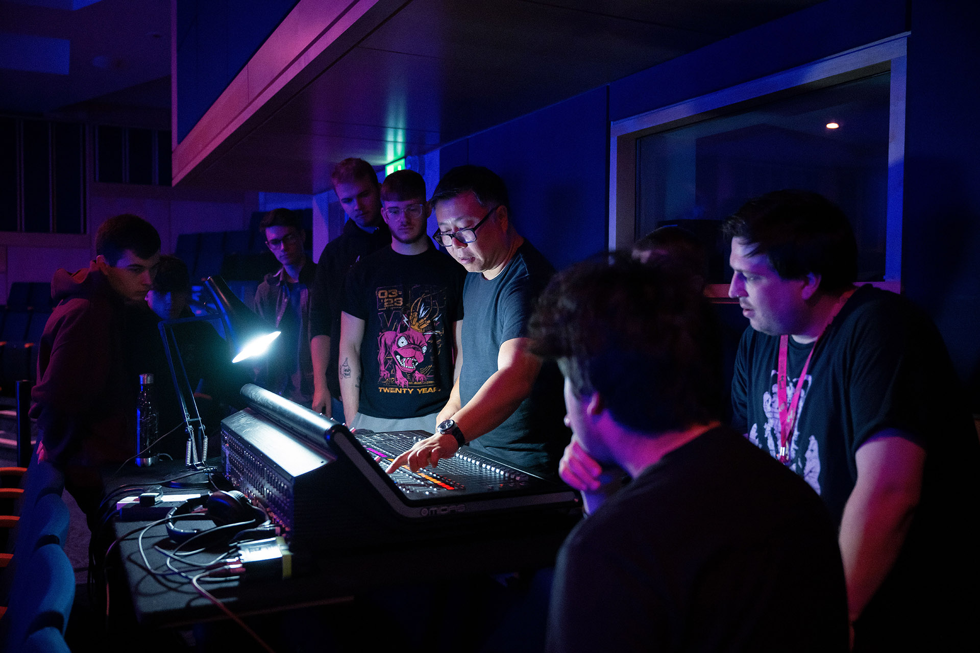 A music lecturer shows functionality of a mixing desk to a group of students crowded round