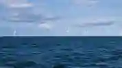 Numerous offshore wind turbines on the horizon under a cloudy blue sky with a choppy sea in the foreground.