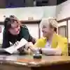 Two actors sat leaning over a table on stage.