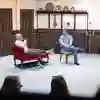 Two actors sat in armchairs on stage.