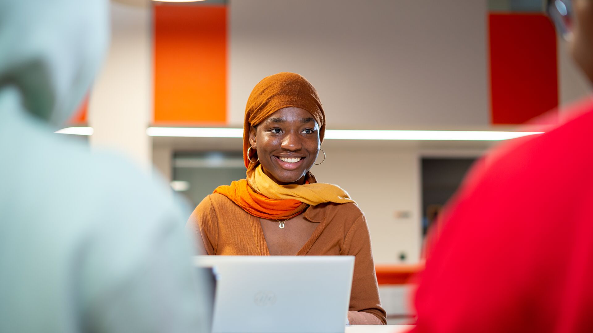 Female student in front of a laptop talking to 2 others seen in image from behind