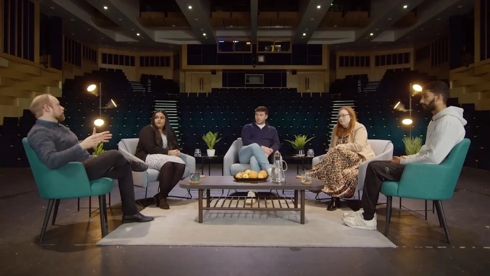 A group of people sit in comfortable chairs around a coffee table on a stage