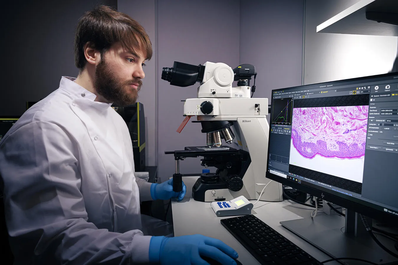 A researcher in white coat sits in front of a microscope and a computer screen showing a close up of wound tissue