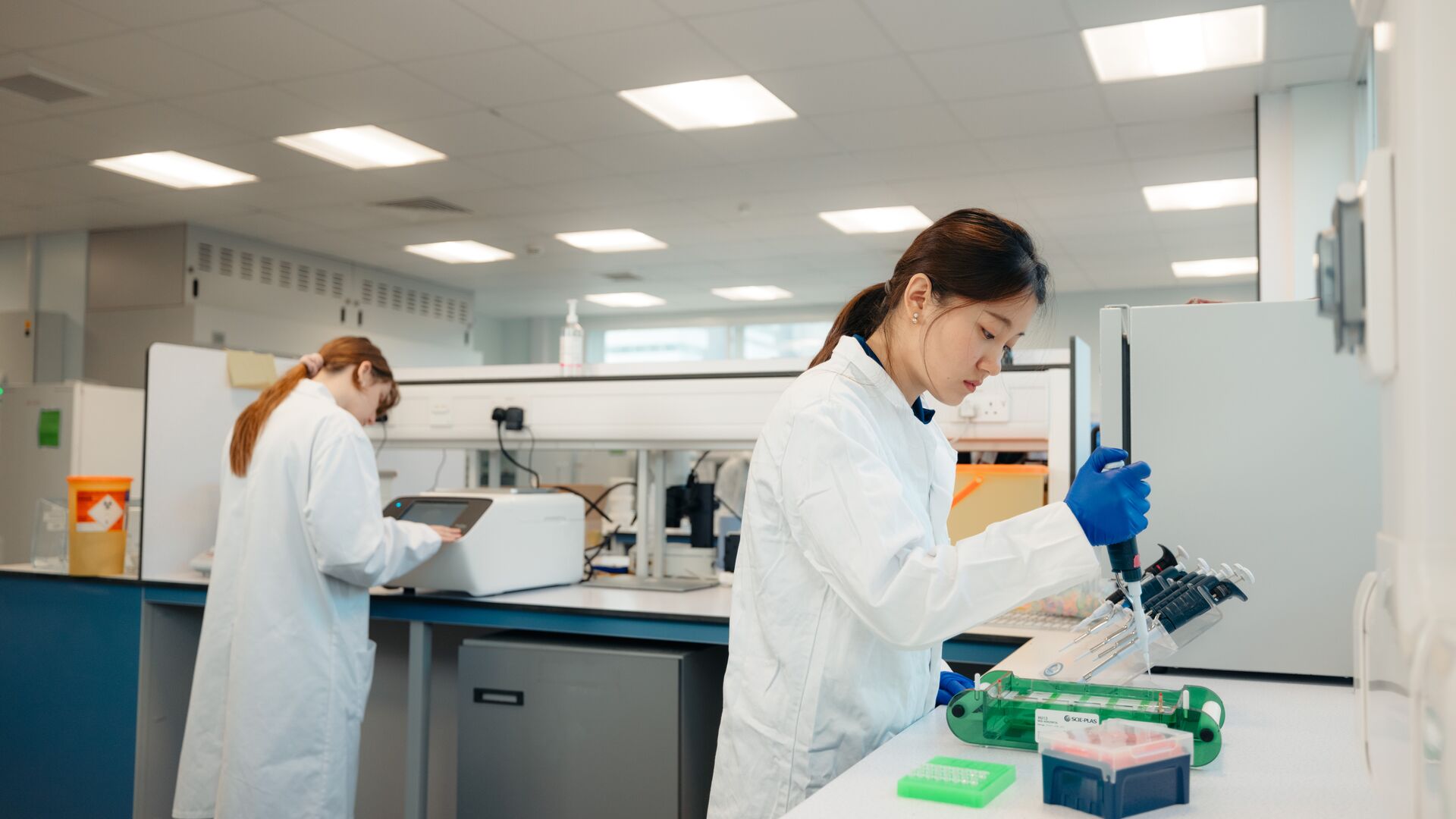 A student in white lab coat uses a pipette at a lab bench