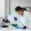 Female student in a white lab coat observes an item intently, surrounded by laboratory equipment.