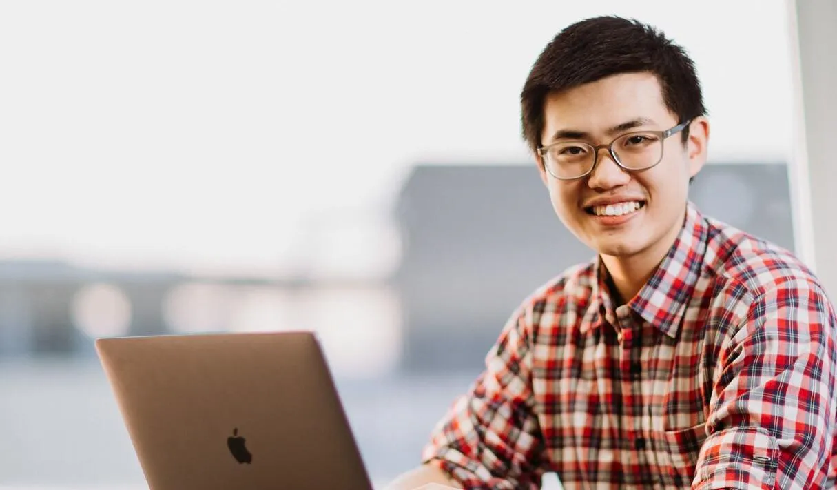 Student in library with laptop, smiling at the camera.