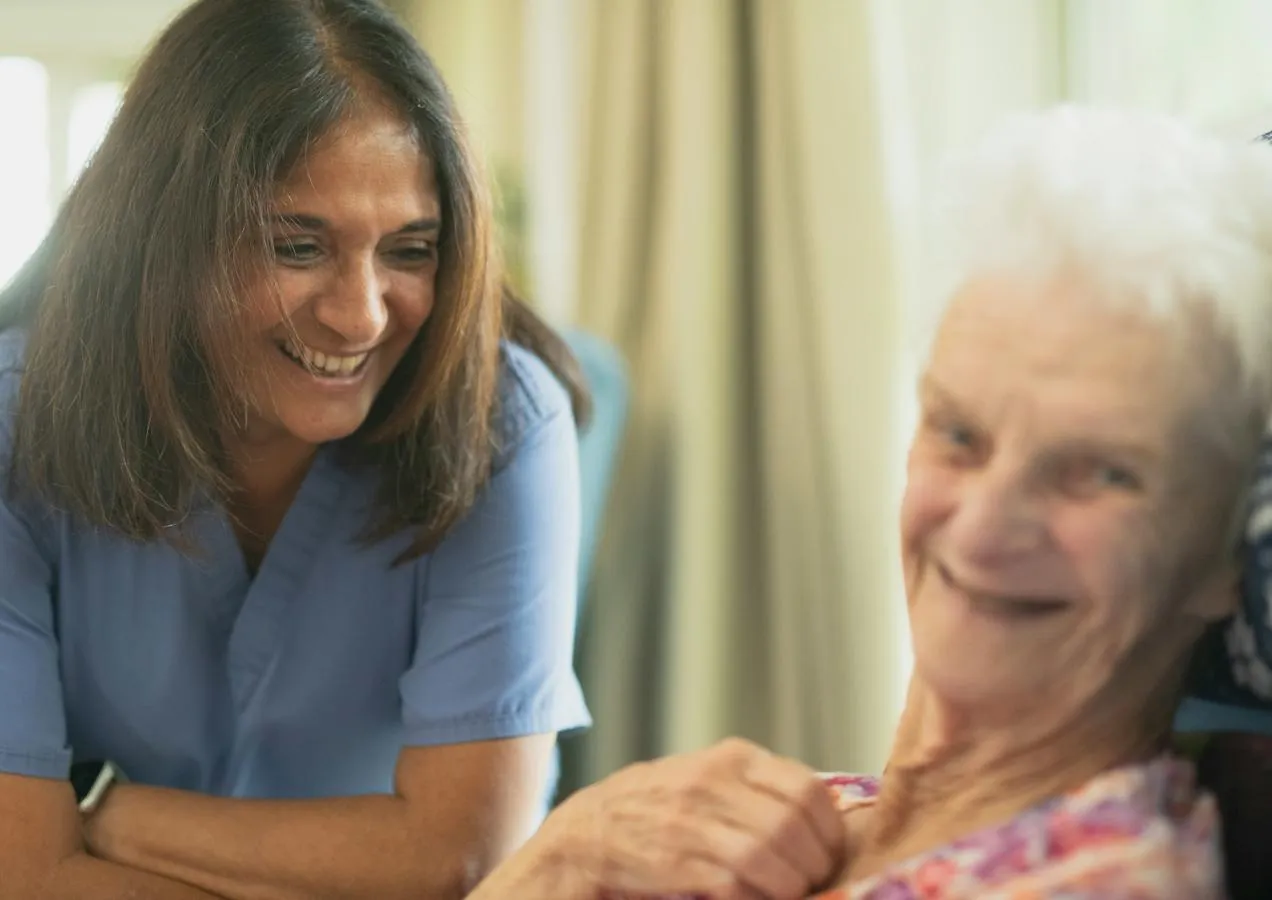 An older woman is smiling at the camera with a smiling carer at her side