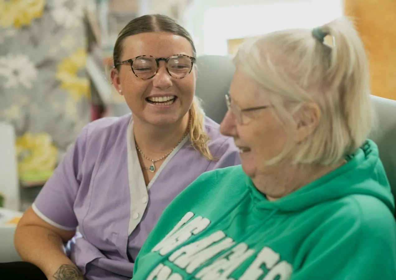 a younger woman in carers uniform and older woman sitting side by side share a laugh 
