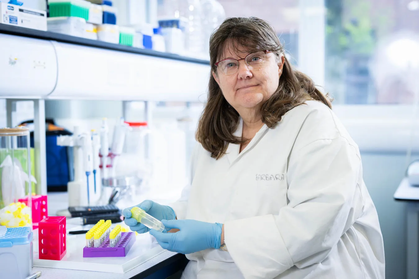 woman in white lab coat seated at lab bench holding a sample tube.