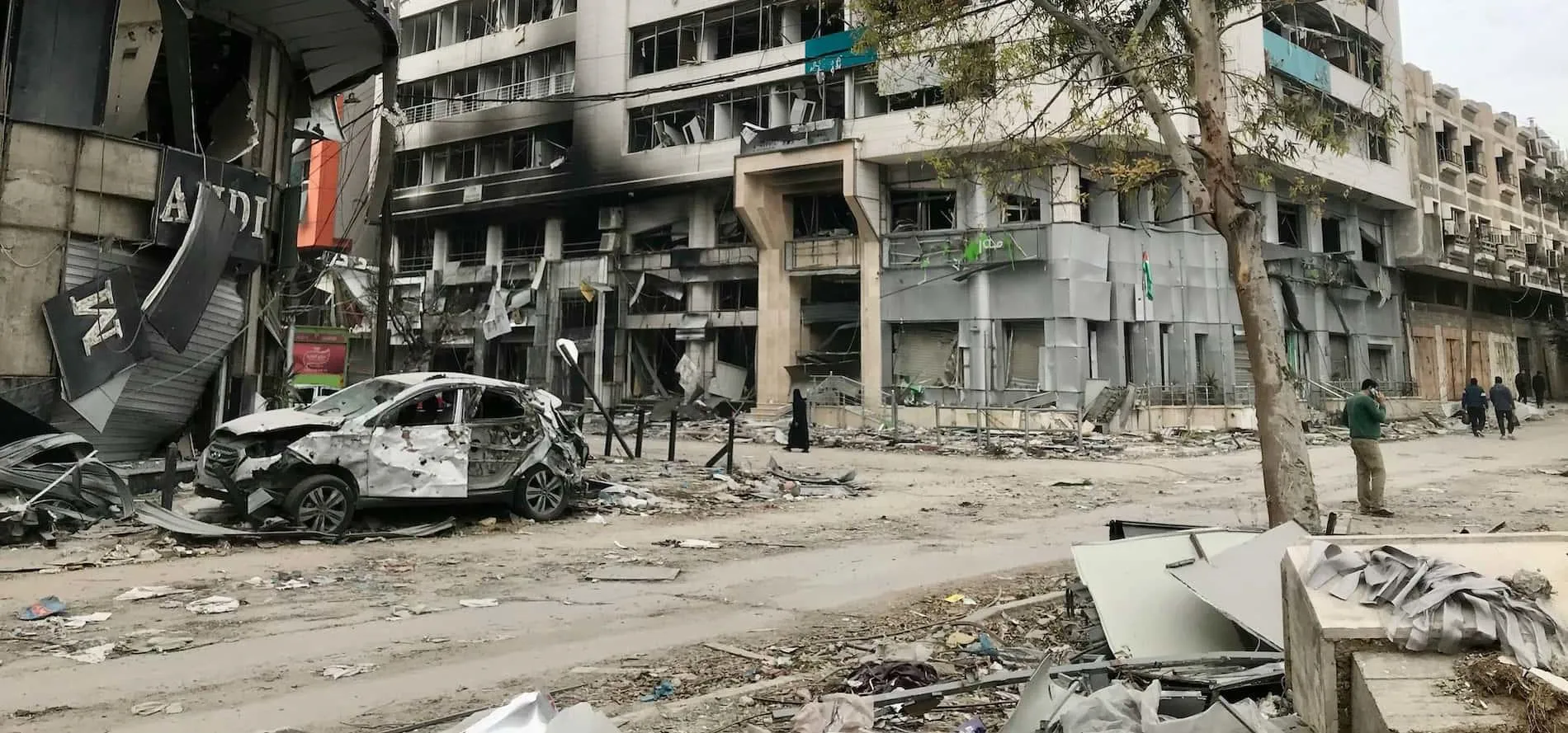 a bombed out out car parked at the side of a city street filled with charred and blasted buildings