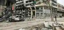a bombed out out car parked at the side of a city street filled with charred and blasted buildings