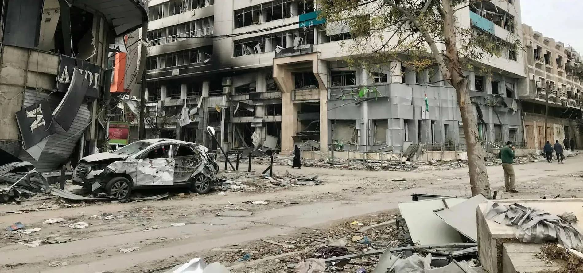 a bombed out out car parked at the side of a city street filled with charred and blasted buildings