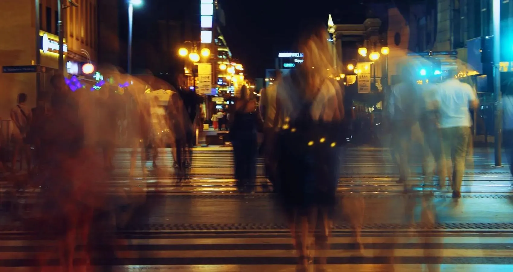 A city centre street at night, blurred people walking over wet roads lit by street lamps and neon signs