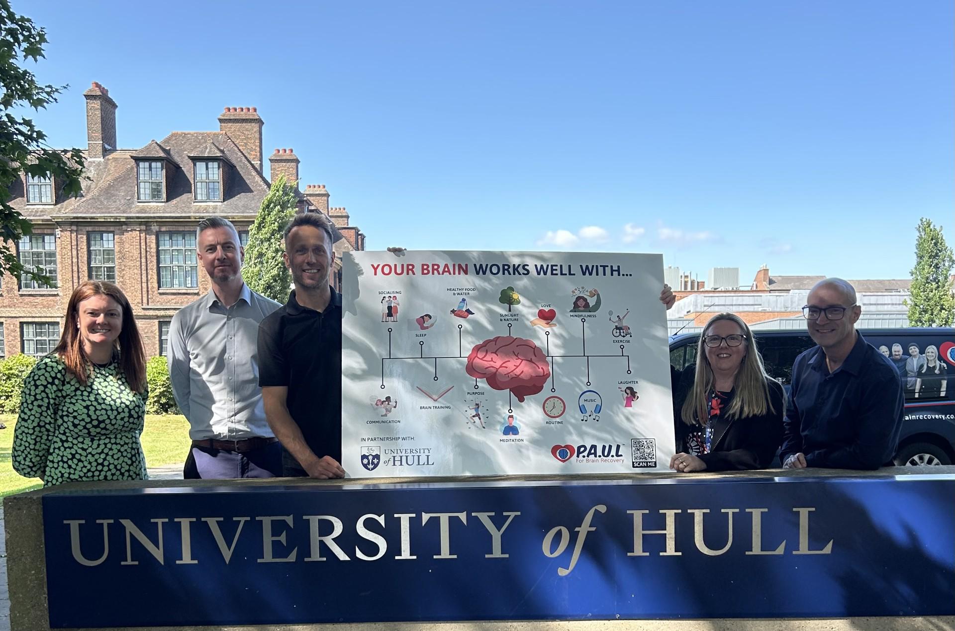 An innovative partnership: Joanne Black (Dietetics) Leigh North, Paul Spence, (P.A.U.L. For Brain Recovery), Dr Kelly Dockerty, Professor Jonathan Glazzard (School of Education) Five people at the entrance to University of Hull holding a Brain Health Board