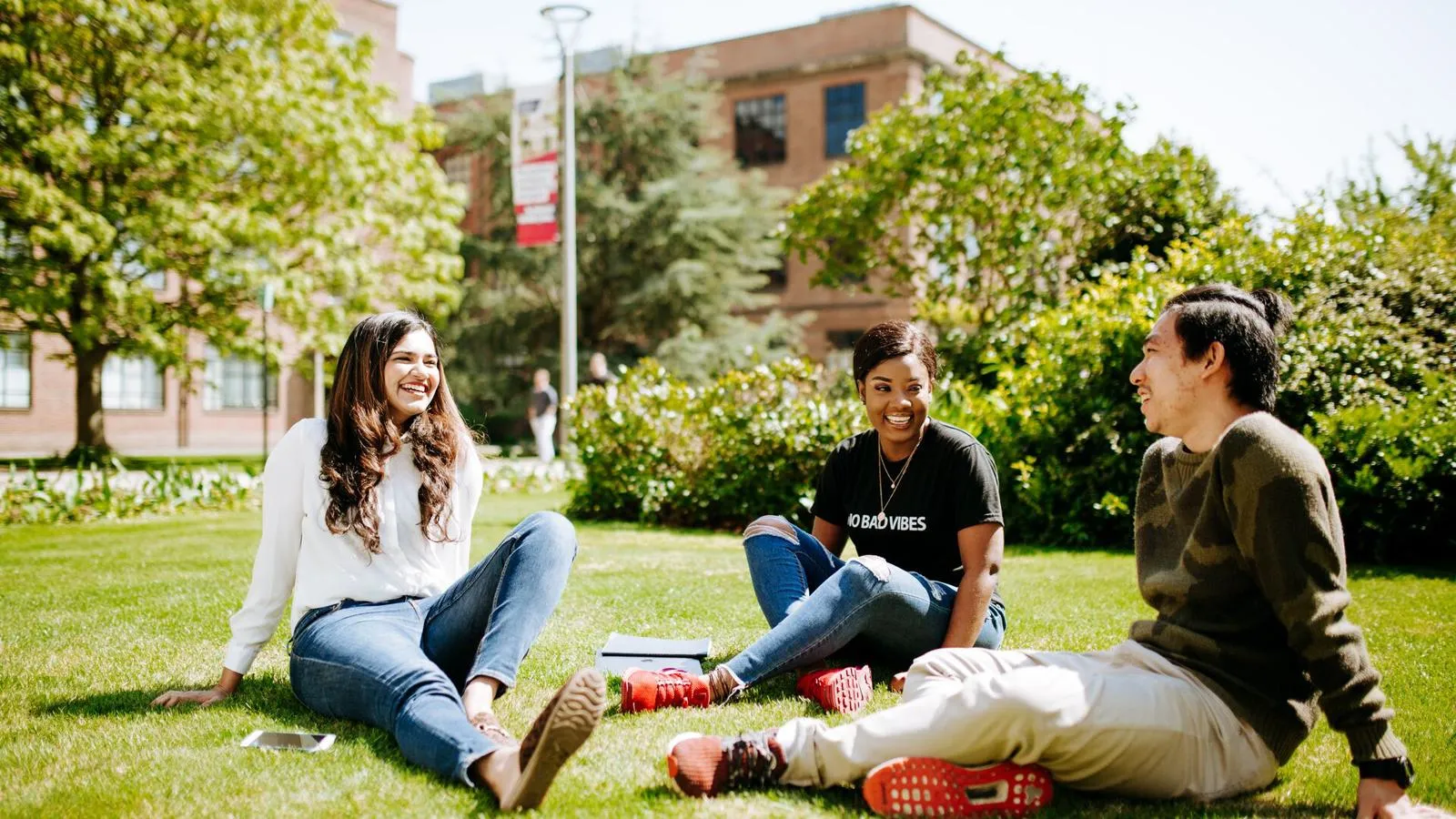 Three people sit on grass in a park-like setting, talking and smiling with trees and a building in the background.