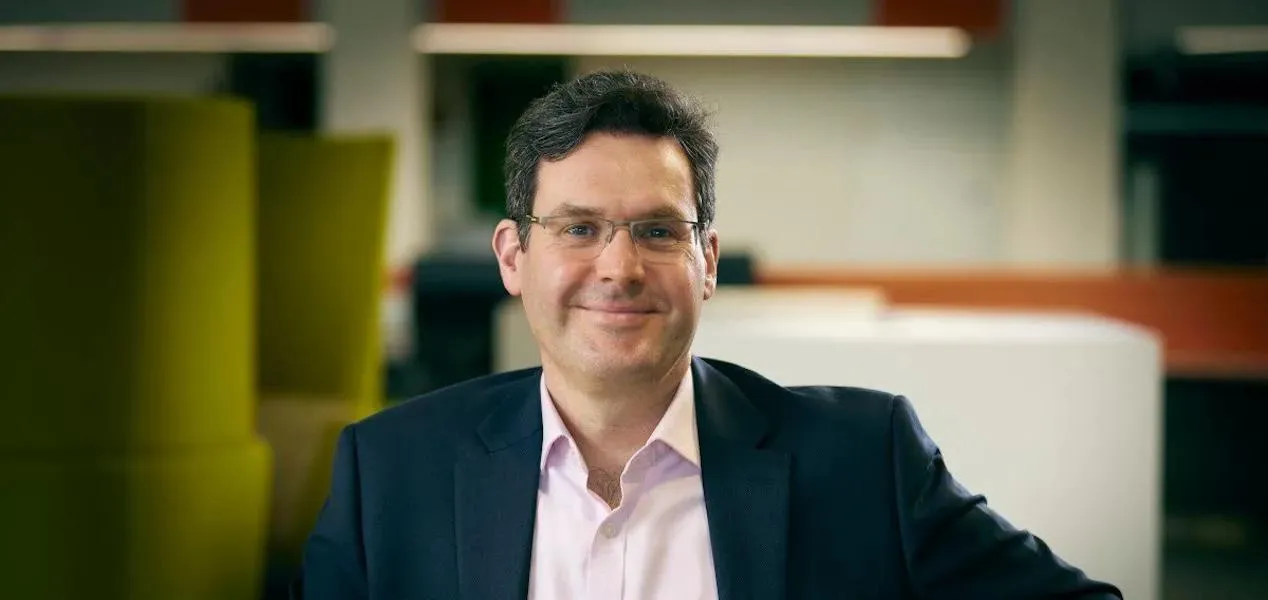 head and shoulders shot of dark haired. bespectacled man in dark suit and pink shirt smiling at camera