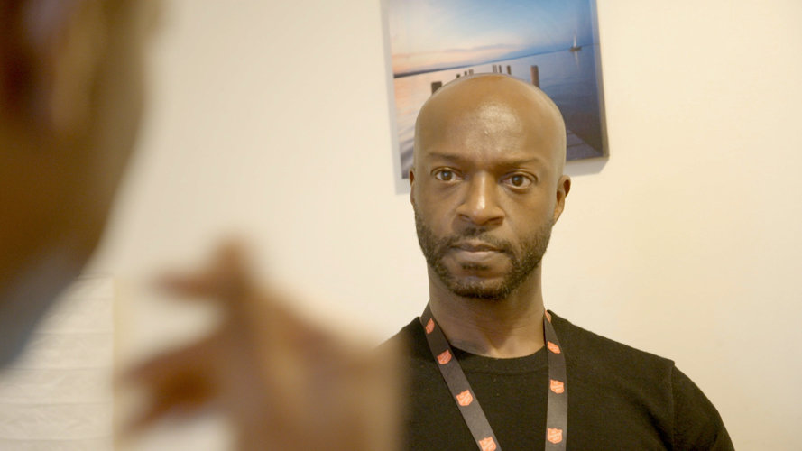 The partnership will tackle some of the most pressing issues faced by victims and practitioners in new innovative and collaborative ways. Picture credit: The Salvation Army. a seated black man wearing a lanyard and black tee shirt facing someone out of focus who is talking to him