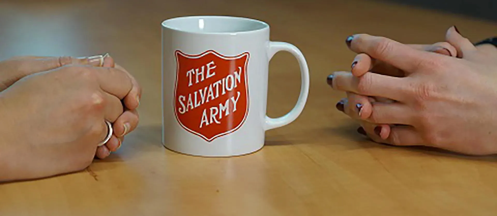 a cup on a table with The Salvation Army written on it and two pairs of hands to either side