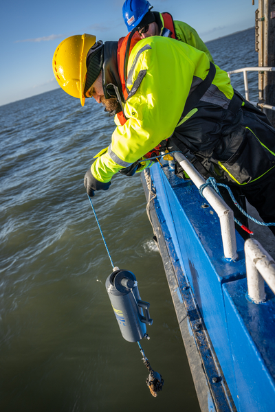 man in hard hat and high viz clothing leaning over the side of a boat and lifting a flask from the sea