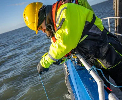 man in hard hat and high viz clothing leaning over the side of a boat and lifting a flask from the sea