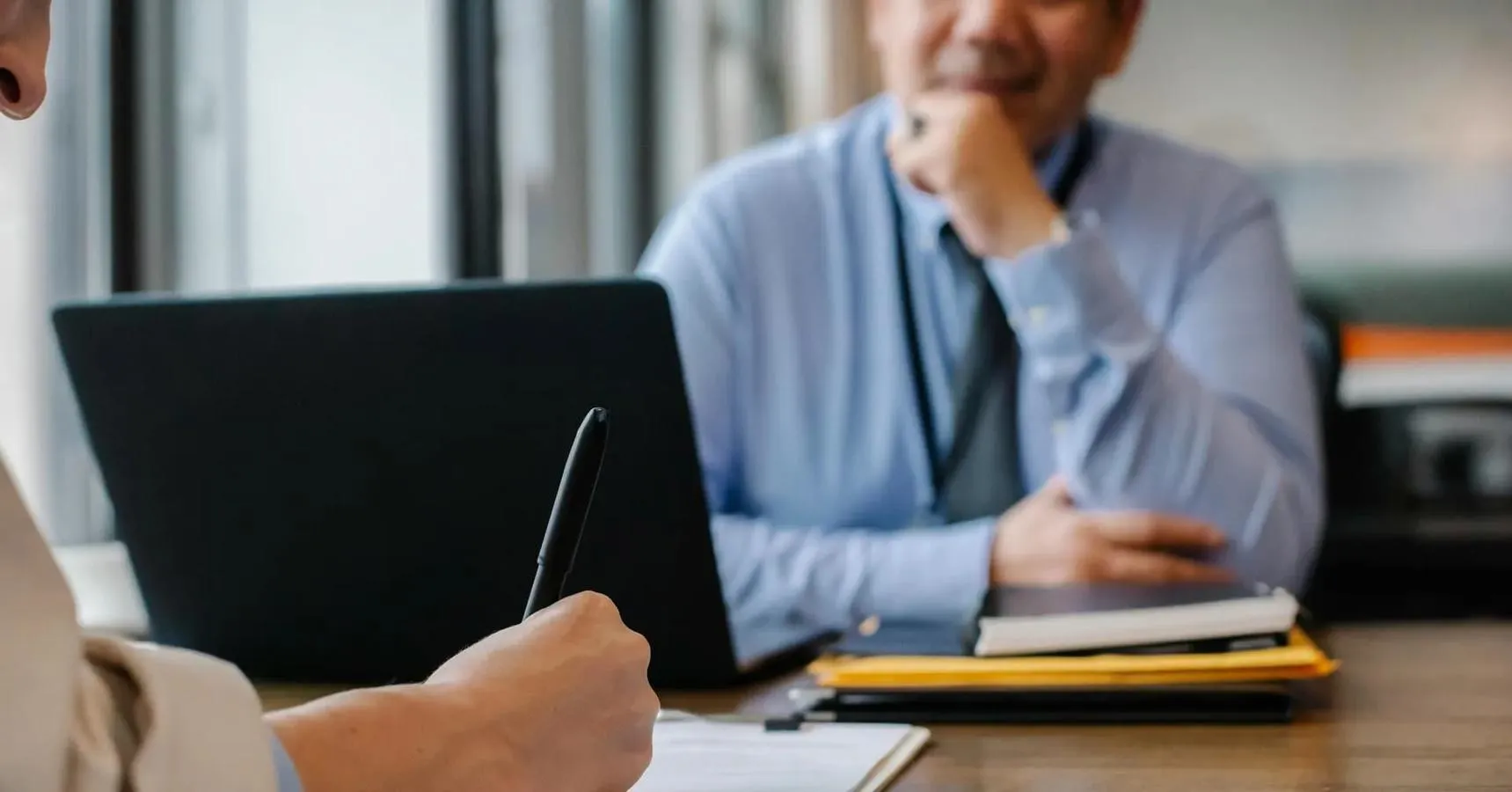 Person writing on paper sat in front of someone on a laptop